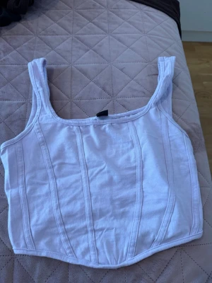 Ljuslila croppad korsettopp - Trendig ljuslila croppad korsettopp från Bershka med breda axelband och markerade sömmar framtill. Toppen har en tight passform och är tillverkad i mjuk bomull med lite stretch. Perfekt för att lyfta din outfit med en modern touch.