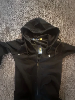 (KIDS) black zipper Ralph Lauren - Svart hoodie från Polo Ralph Lauren med dragkedja framtill, huva med snörning och klassisk logga broderad på bröstet. Tillverkad i mjuk bomull och perfekt för en chill och stilren look. Storlek M för barn.