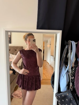 Glittrig vinröd kort klänning - Glittrig vinröd klänning med kort kjol och ärmlös design. Klänningen har en rund halsringning och djup rygg, perfekt för dig som gillar att sticka ut. Materialet är troligtvis polyester med paljetter som ger extra shimmer. Storlek 170 aldrig använd!