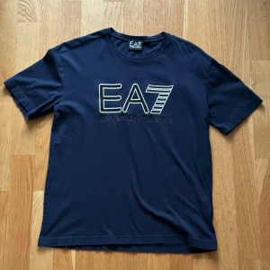 Armani T-Shirt - EA7 Emporio Armani T-shirt i mörkblå färg med stor logga framtill. Stilren, sportig och lättmatchad. Perfekt till vardags eller casual outfits.