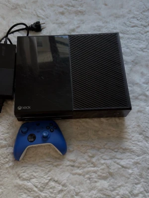 Xbox One med blå handkontroll - Säljer en Xbox One spelkonsol i svart med en snygg blå trådlös handkontroll. Konsolen har en stilren, rektangulär design i plast och kommer med strömadapter. Perfekt för gaming och streaming.