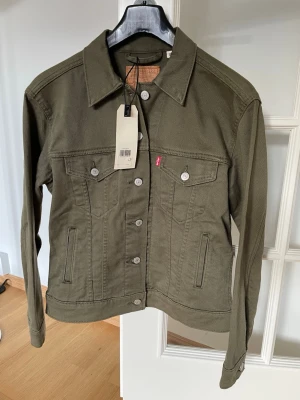 Exboyfriend trucker jacka från Levi's - Stilren olivgrön jeansjacka från Levi's med klassisk krage, silverfärgade knappar och två bröstfickor med lock. Jackan har raka ärmar och diskret röd Levi's-tagg på bröstet. Perfekt för dig som gillar en tidlös look.