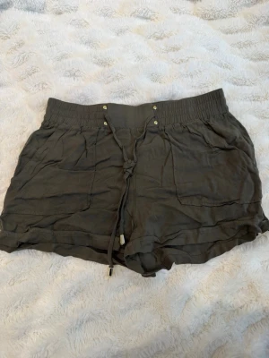 Mörkgröna shorts med snörning - Snygga mörkgröna shorts med bred resår i midjan och snörning framtill. Shortsen har två stora fickor fram och är tillverkade i ett mjukt tyg som känns skönt mot huden. Perfekta för varma dagar och enkel att matcha med olika toppar.
