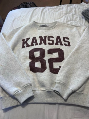 Grå sweatshirt med tryck KANSAS 82 - Snygg ljusgrå sweatshirt med mörkrött tryck 'KANSAS 82' på bröstet. Tröjan har rund hals, ribbade muddar och avslappnad passform. Perfekt för en sportig och avslappnad stil.