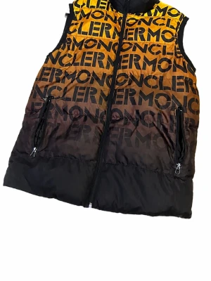 Moncler dunväst med logotypmönster - (kolla 3dje bilden)Snygg dunväst från Moncler i svart och gul-orange gradient med stort logotypmönster över hela västen. Västen har hög krage, två dragkedjeförsedda fickor och hel dragkedja framtill. Perfekt för dig som vill sticka ut med en trendig och exklusiv look.