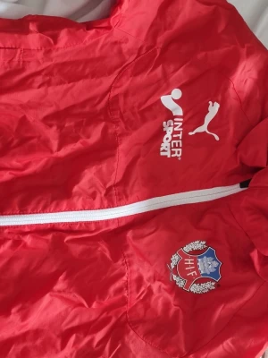 Röd Helsingborgs IF jacka Puma - Röd sportjacka från Puma med Helsingborgs IF klubbmärke på bröstet. Jackan har vit dragkedja och tryckta loggor från Intersport och Puma. Perfekt för fotbollsträning eller som supporterplagg.