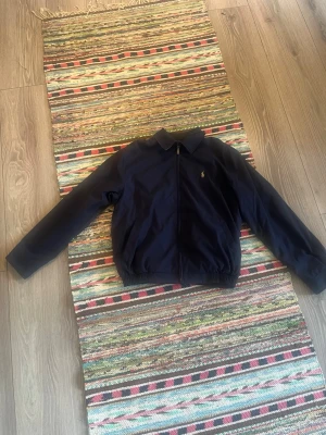 Mörkblå jacka från Ralph Lauren - Snygg mörkblå jacka från Ralph Lauren med dragkedja framtill och klassisk krage. Jackan har elastisk midja och ärmar samt en liten broderad logga på bröstet. Perfekt för dig som gillar stilren och tidlös design.