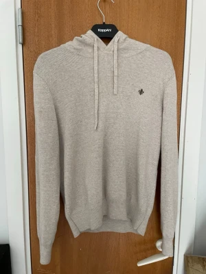 Beige stickad hoodie från KappAhl - Snygg beige stickad hoodie från Morris med dragsko i huvan och diskret broderad logga på bröstet. Tillverkad i mjukt, ribbstickat material som ger en clean och stilren look. Perfekt för dig som gillar enkel och bekväm stil. Hoodien är ganska kort och passar dig som e 165-175