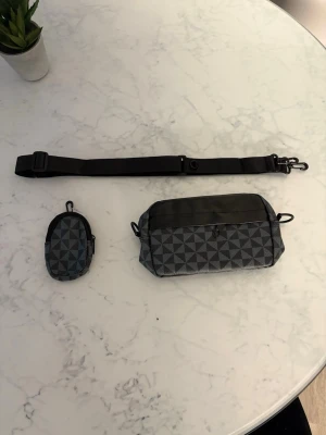 Gråmönstrad midjeväska med liten pouch - Säljer en grå och svart midjeväska med geometriskt mönster och tillhörande liten pouch. Väskan har justerbart och avtagbart band samt dragkedja upptill. Perfekt för att bära småprylar och accessoarer.