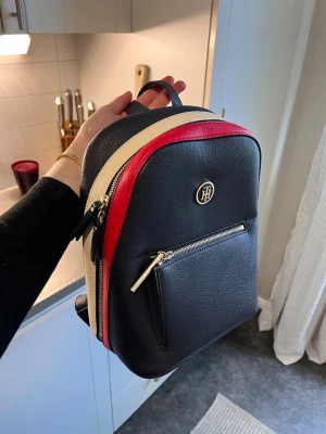 Tommy Hilfiger ryggsäck i skinn - Snygg ryggsäck från Tommy Hilfiger i marinblått skinn med röda och vita detaljer. Guldiga dragkedjor och TH-logga framtill. Praktiskt ytterfack och justerbara axelband. Perfekt för dig som vill ha en stilren och trendig väska. Köpt i Kitzbühl för 4000kr(tror jag)💕