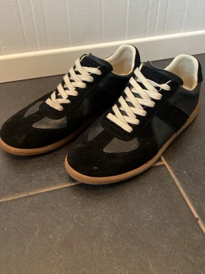 Maison margiela - Snygga svarta sneakers med vita snören och bruna gummisulor. Skorna har en mix av mocka och slätt material på ovandelen samt en klassisk låg siluett. Perfekta för dig som gillar stilrena och tidlösa sneakers.