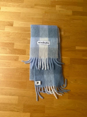 Blå-rutig halsduk från Acne Studios - Supermjuk och fluffig halsduk från Acne Studios i blått och vitt rutmönster. Halsduken har långa fransar i ändarna och stor logotyp framtill. Perfekt accessoar för att lyfta din outfit med en trendig touch.
