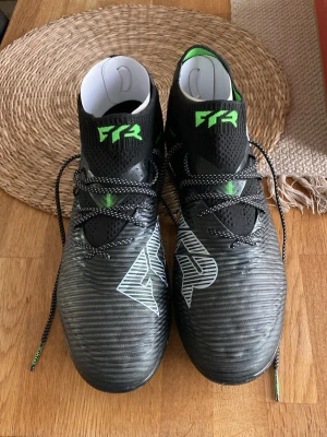 Svarta Puma fotbollsskor med gröna detaljer - Säljer ett par svarta fotbollsskor från Puma med gröna och vita detaljer. Skorna har snörning, hög krage och mönstrad ovansida för bättre bollkontroll. Perfekta för dig som vill ha stil och funktion på planen. Använda en gång på en träning då han insåg att de var lite för stora. 