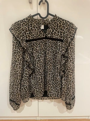 Leopardmönstrad blus med volanger - Trendig blus med leopardmönster och svarta volangdetaljer längs axlar och ärmar. Blusen har hög hals och långa ärmar med svarta band vid ärmslut. Perfekt för dig som vill sticka ut med en cool och djärv stil.