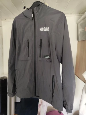 Grå vindjacka från Hoodrich - Snygg grå vindjacka från Hoodrich med huva och justerbara snören. Jackan har flera dragkedjeförsedda fickor på bröstet och sidorna samt vit logga på bröstet. Perfekt för streetwear-stil och skyddar mot vind.