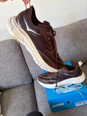 U Hoka Mafate Speed 4 lite - Otroligt lätta och snygga bruna Hoka Mafate Speed 4 sneakers med vita detaljer och tjock, krämfärgad sula. Ovandelen är i mesh och syntetmaterial med reflekterande detaljer. 