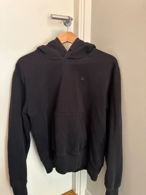 Svart hoodie från Acne Studios - Säljer en svart hoodie från Acne Studios med klassisk huva och magficka. Tröjan har en liten fyrkantig logotyp på bröstet och är tillverkad i mjuk bomull. Ett litet hål på högra axeln, ca en halv cm. 