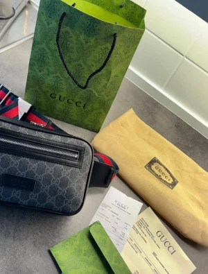 Gucci axelväska med monogram - Snygg axelväska från Gucci i svart canvas med klassiskt GG-monogram och svarta läderdetaljer. Väskan har en framficka med dragkedja och justerbar axelrem i rött och blått. Perfekt för dig som vill ha en trendig och lyxig accessoar.