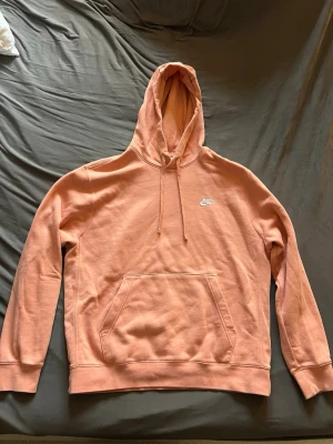 Persikofärgad Nike hoodie - Snygg persikofärgad hoodie från Nike med klassisk logga broderad på bröstet. Hoodien har huva med dragsko, magficka och ribbade muddar vid ärmslut och nederkant. Kom gärna med prisförslag. 