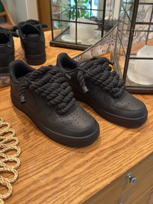 Svarta Nike Air Force 1 med tjocka snören - Säljer ett par helsvarta Nike Air Force 1 sneakers med unika, extra tjocka svarta snören. Skorna har klassisk låg siluett, perforerad tå och är tillverkade i läder med gummisula. Perfekta för dig som vill sticka ut med en fet detalj på fötterna.