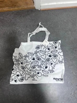Vit shoppingväska från Flying Tiger - Vit shoppingväska i tyg med svarta grafiska tryck av ögon, händer och läppar på ena sidan och ett stort öga på andra sidan. Väskan har två handtag och är perfekt för att bära med sig grejer. Snygg och lekfull design.