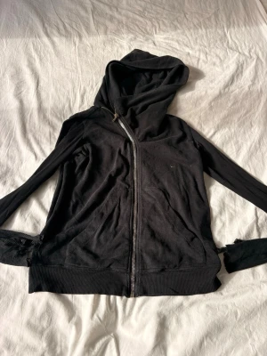 Svart hoodie med sned dragkedja - Svart hoodie med cool sned dragkedja och stor huva. Mjukt bomullsmaterial och två fickor framtill. Perfekt för en avslappnad streetstyle-look. Passar dig som gillar enkla men snygga detaljer.