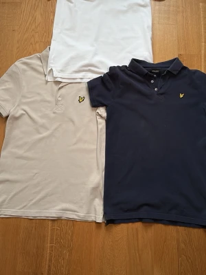 3-pack pikétröjor från Lyle & Scott - Tre klassiska pikétröjor från Lyle & Scott i vitt, beige och marinblått. Alla har korta ärmar, krage, knappar framtill och den gula loggan på bröstet. Perfekt nu till sommaren. Storleken är xs på alla tröjorna och perfekt till ett par jeans eller linnebyxor till sommaren. Väldigt bra skick inte jätte mycket användning har mest hängt i garderoben. 