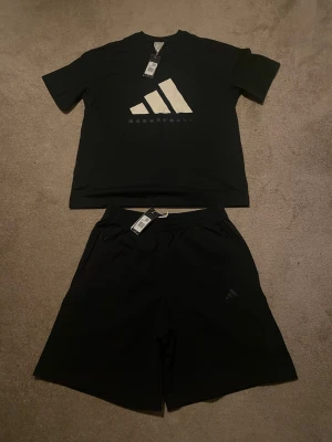 Svart basketset från Adidas - överdel M, underdel L - Snyggt svart basketset från Adidas med t-shirt och shorts. T-shirten har stort vitt Adidas-tryck och texten 'Basketball' framtill. Båda plaggen är i mjukt och bekvämt material, perfekt för träning eller match. Klassisk sportig look med diskreta detaljer.