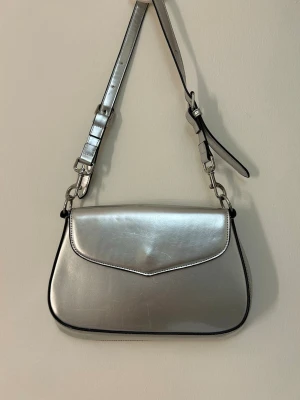 Silvrig Public Desire axelväska  - Snygg axelväska i metallic-silver med justerbar axelrem och stilren design. Väskan har ett lock framtill med svarta kanter och silvriga metalldetaljer. Har en en liten fläck på insidan av väskan och små repor. Skriv om flera bilder önskas 🥰