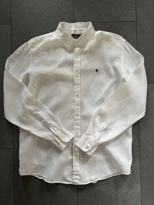 Vit linneskjorta från Morris - Klassisk vit skjorta från Morris i linne med button down-krage och diskret broderad logga på bröstet. Skjortan har långa ärmar och knappar framtill, perfekt för en clean och stilren look. Skjortan är i gott skick men har en liten skada (se sista bild)