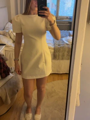 Vit kort klänning med puffärm från Zara  - Snygg vit kort klänning med puffärmar och rund halsringning. Klänningen har en figurnära passform och är perfekt för student eller sommarens tillfällen. Enkel och stilren design utan mönster.