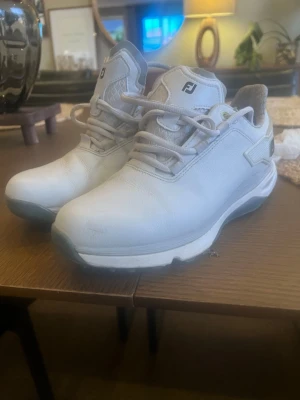 Vita sneakers från FootJoy - Säljer ett par vita sneakers från FootJoy med lågt skaft och snörning. Skorna har en stilren design med grå detaljer och är tillverkade i ett tåligt material som passar perfekt för en sportig look.