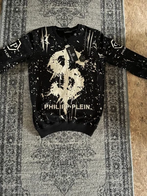 Svart tröja Philipp Plein dollartryck - Cool svart tröja från Philipp Plein med stort dollartryck i vitt framtill och stänkmönster över hela plagget. Långärmad modell med ribbade muddar och logotypdetaljer på ärmarna. Perfekt för dig som gillar streetstyle och statement-plagg.