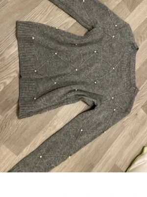 Grå stickad tröja med pärldetaljer - Snygg grå stickad tröja med dekorativa pärlor över hela plagget. Tröjan har lång ärm, rund halsringning och ribbstickade muddar. Perfekt för dig som vill ha en trendig och mysig look med det lilla extra.