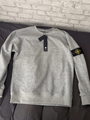 Grå sweatshirt från Stone Island - Snygg grå sweatshirt från Stone Island med klassisk rund hals och ribbade muddar. Tröjan har den ikoniska loggan på vänster ärm och är gjord i mjukt bomullsmaterial. Perfekt för en clean och stilren look.