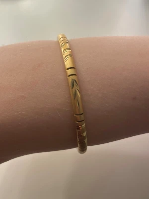 Guldfärgat stelt armband med mönster - Snyggt stelt armband i guldfärg med ingraverade mönster och svarta detaljer. Armbandet har en klassisk rund form och är enkelt att matcha med andra smycken. Perfekt accessoar för att lyfta din stil. Helt oanvänd och ej äkta guld 🫶
