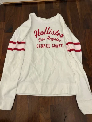 Vit långärmad tröja Hollister - Vit långärmad tröja från Hollister med röd text 'Hollister Los Angeles Sunset Coast' på bröstet och två röda ränder på varje ärm med en ljusrosa rad i mitten. Tröjan har rund halsringning och är i ett mjukt material.