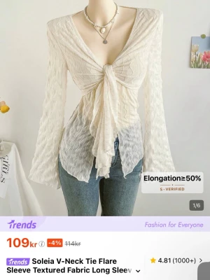 Vit v-ringad blus med knytning - Trendig vit blus från Soleia med v-ringning och knytning framtill. Blusen har långärmade utsvängda ärmar och är tillverkad i ett lätt, texturerat tyg med vågigt mönster. Perfekt för en stilren och modern look.