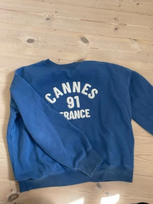 Blå sweatshirt med Cannes-tryck - Cool blå sweatshirt med vitt tryck på ryggen där det står 'Cannes 91 France'. Tröjan har rund hals, ribbade muddar och är gjord i mjukt material. Perfekt för dig som gillar avslappnad stil.