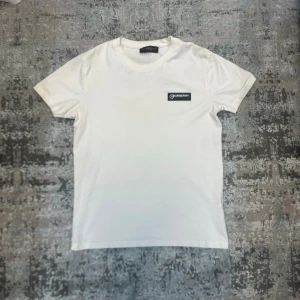 Vit t-shirt - Vit t-shirt med rund halsringning och en liten svart logotyp-patch på bröstet. Klassisk passform och enkel design som passar till det mesta. Tillverkad i mjuk bomull för en bekväm känsla.