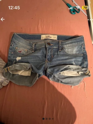 Blå väldigt lågmidjade jeansshorts från Hollister - Snygga blå jeansshorts från Hollister med slitna detaljer och upprullade benslut. Klassisk femficksmodell med låg midja och knappgylf. Perfekta för varma dagar och en avslappnad stil. Byxorna går inte att köpa längre så därav priset men det är diskuterbart. 
