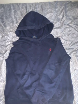 Mörkblå hoodie från Polo Ralph Lauren - Mörkblå hoodie från Polo Ralph Lauren med klassisk huva och den ikoniska röda broderade loggan på bröstet. Mjuk bomullskänsla och avslappnad passform, perfekt för en chill och stilren look.