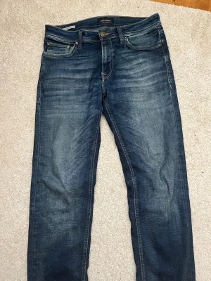 Blå jeans från Jack & Jones Regular W29 L32 - Snygga blå jeans från Jack & Jones i modellen Regular/Clark. Jeansen har klassisk femficksdesign, normal passform och subtila slitningar framtill för en cool look. Perfekta till vardagsstilen och lätta att matcha med olika outfits.