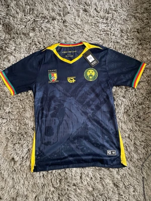 Cameroun fotbollströja navy Fourteen - Snygg navyblå fotbollströja från Fourteen med Camerouns landslagslogga och lejonmotiv på bröstet. Gula detaljer vid krage och sidor, samt röd, gul och grön rand på ärmsluten. Perfekt för dig som älskar fotboll och vill sticka ut på planen.