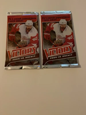 Victory 2009-10 hockeykortspaket - Två oöppnade paket Victory 2009-10 hockeykort från Upper Deck. Varje paket innehåller 6 kort och har en röd och silverfärgad design med hockeyspelare på framsidan. Perfekt för samlare eller hockeyfans som vill hitta svenska superstjärnor.