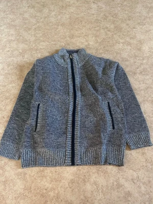 Grå stickad kofta med fleecefoder - Mysig grå stickad kofta med dragkedja och två fickor framtill. Insidan är fodrad med mjuk fleece i rutigt mönster i blått, vitt och brunt. Perfekt för kyliga dagar och enkel att matcha med jeans eller chinos.