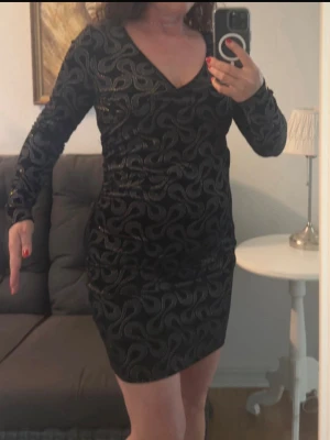 Svart kort klänning med glittrigt mönster - Svart klänning med v-ringning och långärmad design. Klänningen har ett glittrigt mönster som ger en cool look och är figurnära med kort längd. Perfekt för dig som gillar att sticka ut. Materialet känns mjukt och stretchigt, troligen polyester.