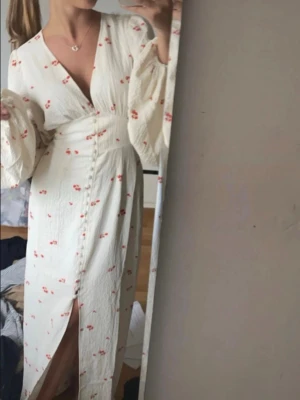 Adoore Vit blommig långklänning med slits - Romantisk vit långklänning med små röda blommor, v-ringning och långa vida ärmar. Klänningen har en slits framtill och markerad midja för en feminin siluett. Perfekt för varma dagar och somriga tillfällen.