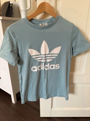 Ljusblå Adidas t-shirt med logga - Snygg ljusblå t-shirt från Adidas med den klassiska vita trefoil-loggan tryckt framtill. T-shirten har rund halsringning och korta ärmar. Tillverkad i mjuk bomull för skön känsla och avslappnad passform.