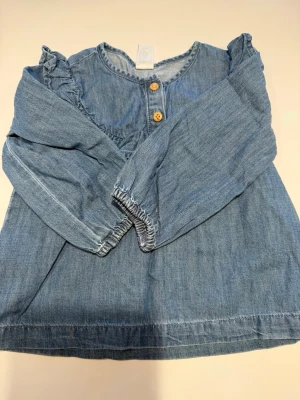 Blå jeansblus med volangdetaljer - Snygg blå jeansblus i denim med volangdetaljer på axlarna och två träknappar framtill. Blusen har rund halsringning och långa ärmar med resår vid ärmslut. Perfekt för en avslappnad och trendig look.